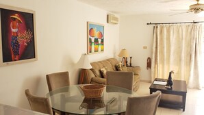 A living room with a glass coffee table, beige sofa, and a dining table with chairs. There are two paintings on the wall, a ceiling fan, and a window with curtains. - Villa 410D at Jolly Harbour (Jolly Harbour)