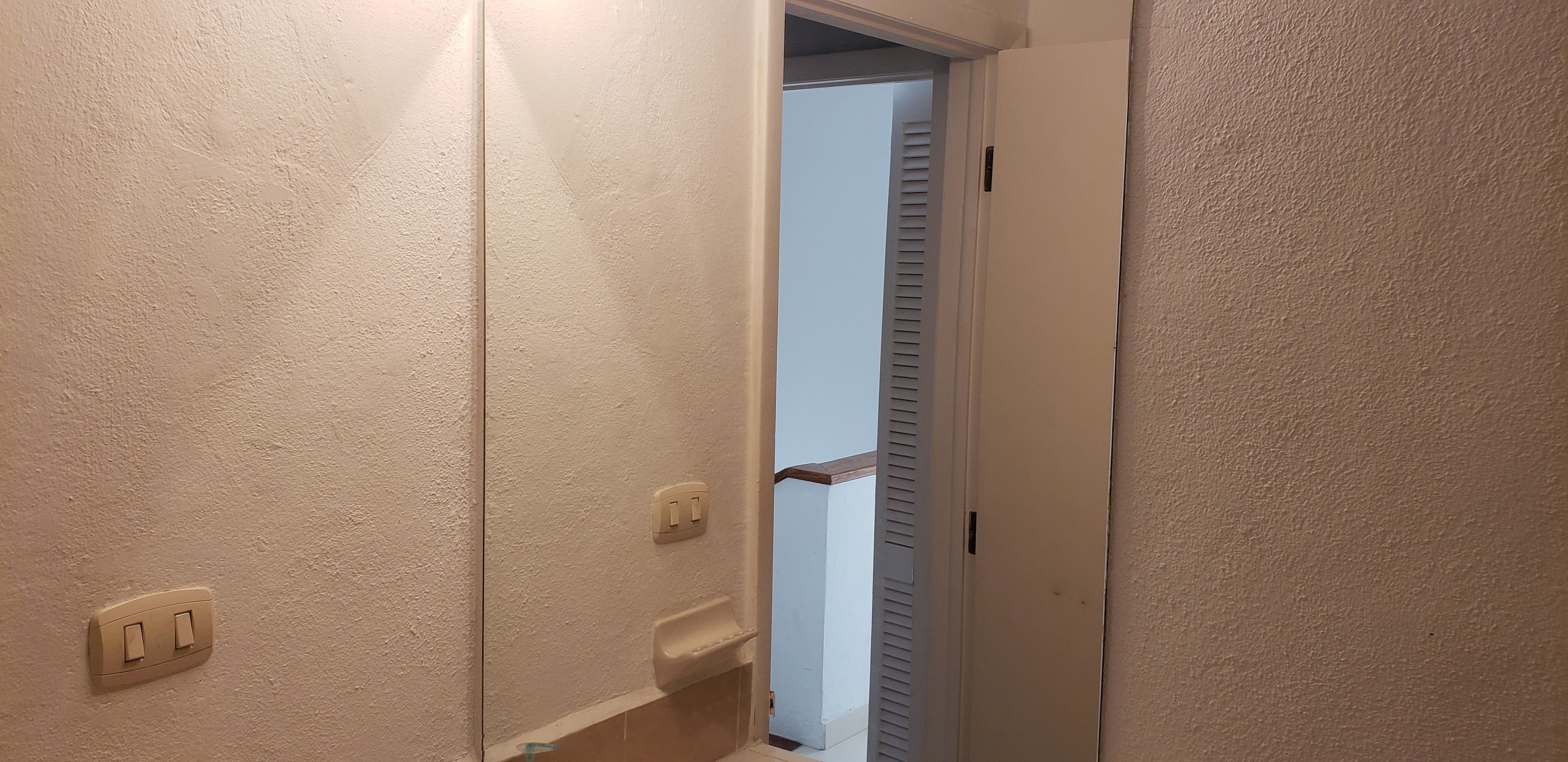 Un pasillo estrecho con paredes beige, una puerta con ventanilla persiana y un interruptor de luz en la pared.