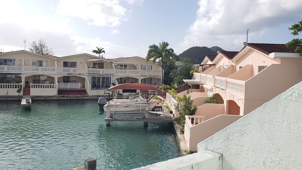 A waterfront hotel with multiple balconies, a dock with a boat, and a mountain in the background. - Villa 410D at Jolly Harbour (Jolly Harbour)