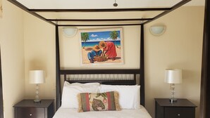 A hotel room with a four-poster bed, two bedside lamps, a framed beach painting, and a floral throw pillow. - Villa 410D at Jolly Harbour (Jolly Harbour)
