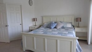 A bedroom with a white four-poster bed, two bedside tables with lamps, a clock on the wall, and a door. - Villa 244D at Jolly Harbour (Jolly Harbour)