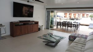 A modern living room with a glass coffee table, a sofa, a TV mounted on the wall, and a sliding glass door leading to an outdoor dining area. - Villa 227E at Jolly Harbour-Adults Only (Jolly Harbour)