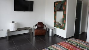 A hotel room with a flat-screen TV mounted on the wall, a bench with a trash bin, a brown leather chair, and a colorful rug. - Villa 227E at Jolly Harbour-Adults Only (Jolly Harbour)