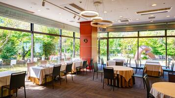 Un restaurante con varias mesas y sillas, amplias ventanas que ofrecen vista a follaje, y luminarias modernas.