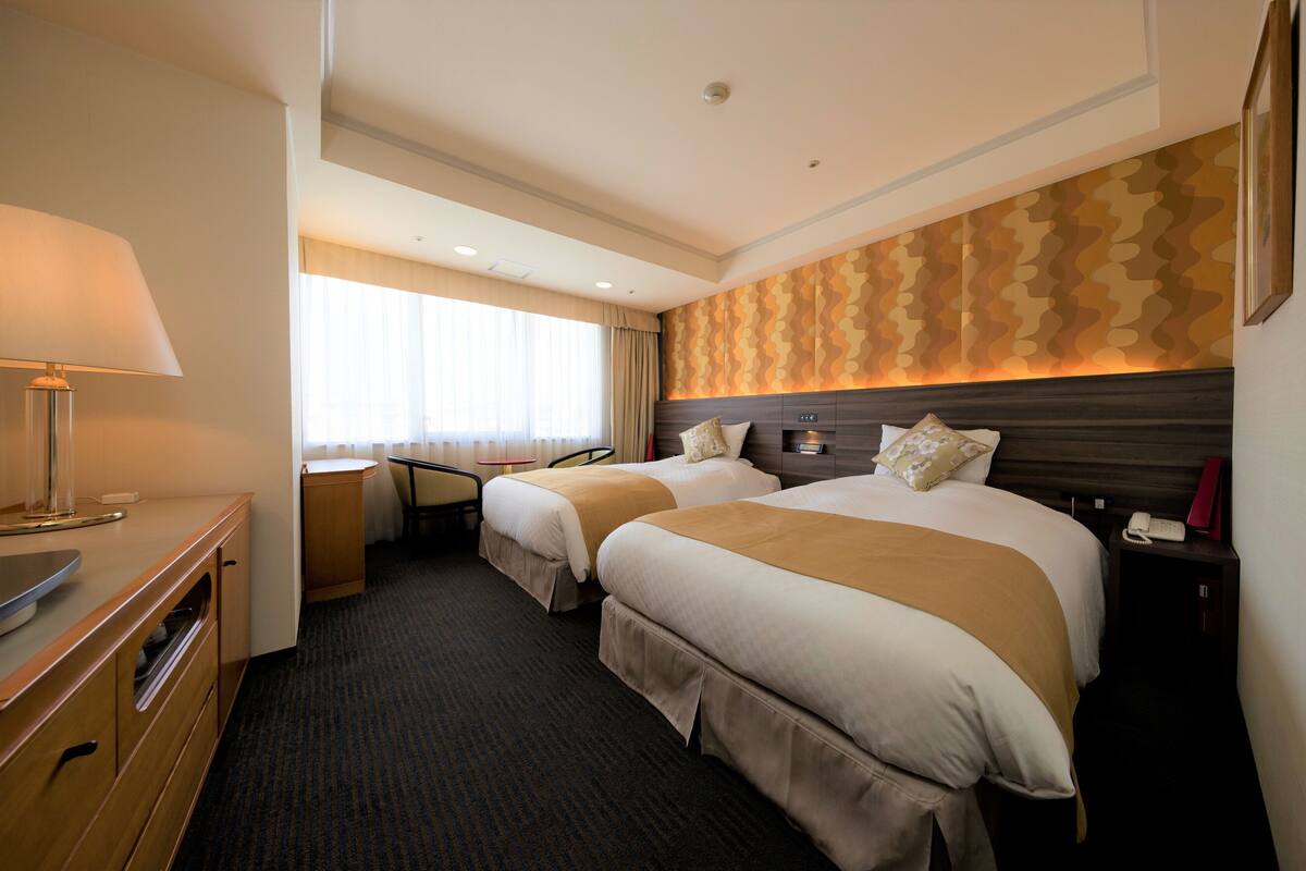 A hotel room with two beds, a desk, a lamp, and a window with curtains.