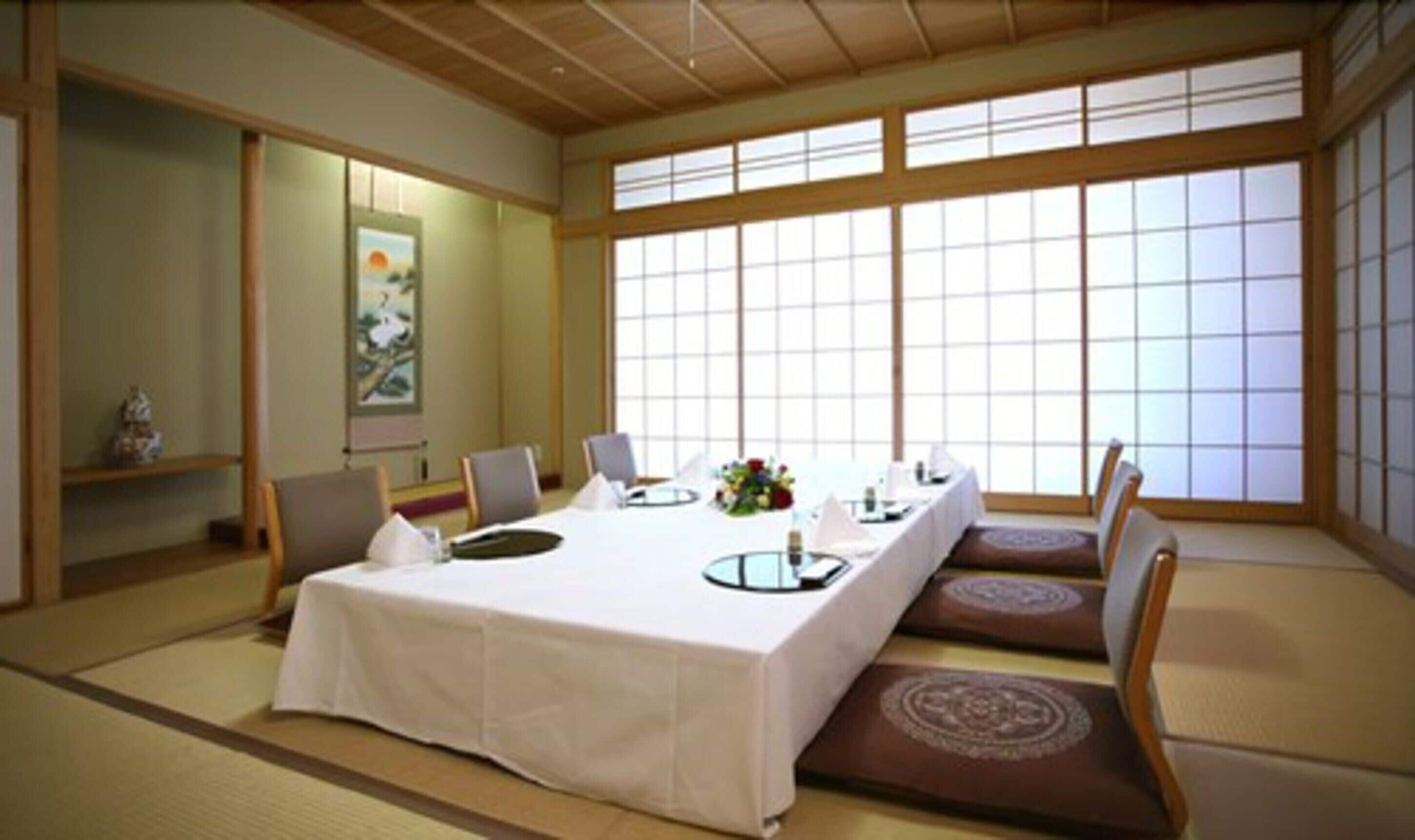 Una habitación japonesa tradicional con suelo de tatami, una mesa baja con mantel blanco y un amplio ventanal con celosías shoji.