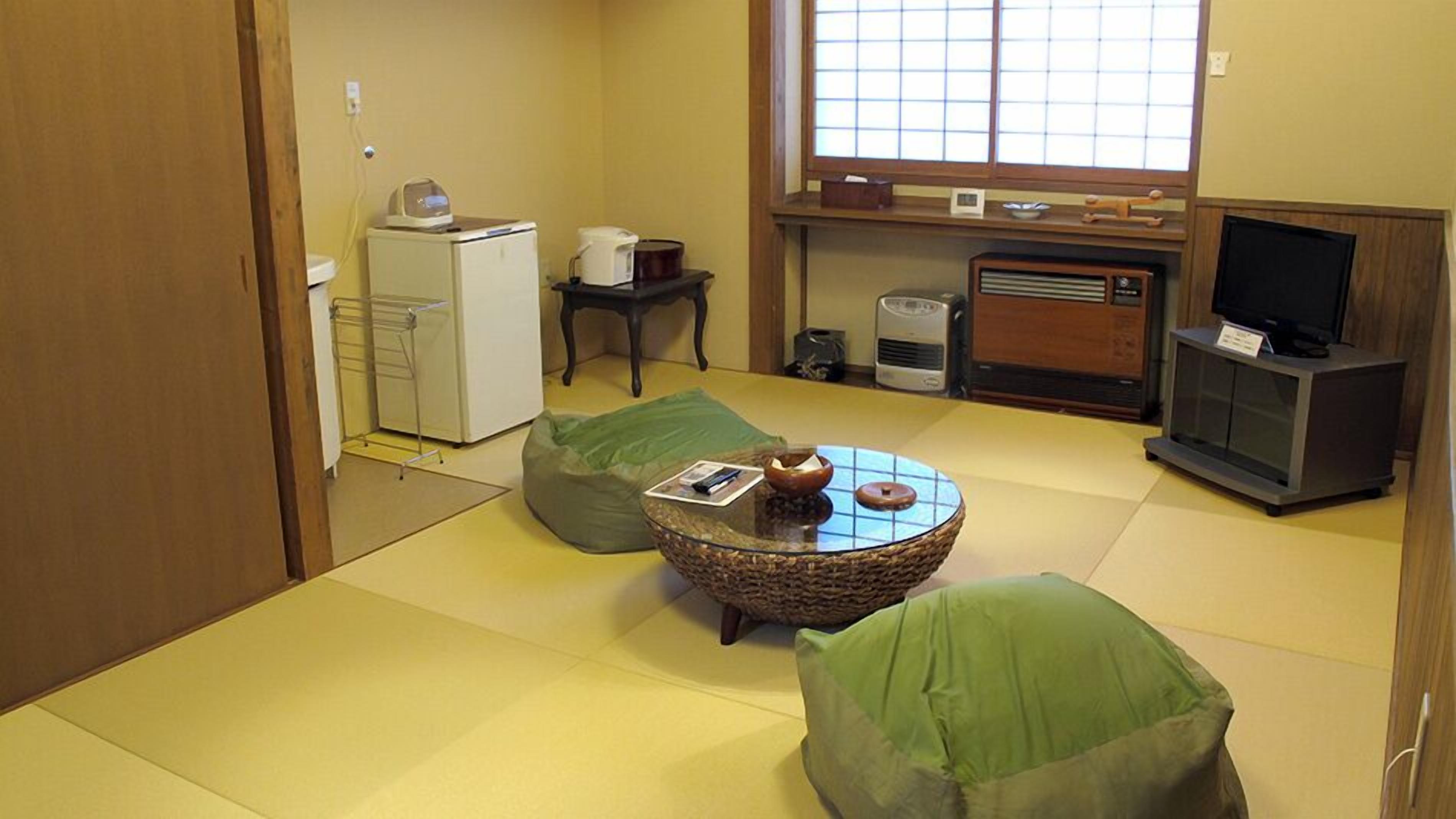 Une pièce de style japonais traditionnel, avec un sol recouvert de tatamis, une petite table basse dont le plateau est fait de fibres tressées, des coussins verts, un téléviseur et un petit réfrigérateur.