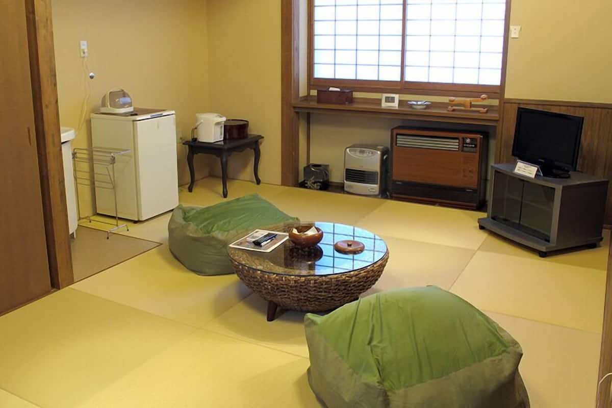 Une pièce de style japonais traditionnel, avec un sol recouvert de tatamis, une petite table basse dont le plateau est fait de fibres tressées, des coussins verts, un téléviseur et un petit réfrigérateur.
