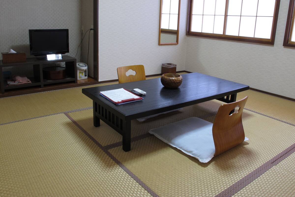 Ein traditioneller japanischer Raum mit Tatami-Boden, einem niedrigen Tisch und einem Fernseher auf einem Regal.