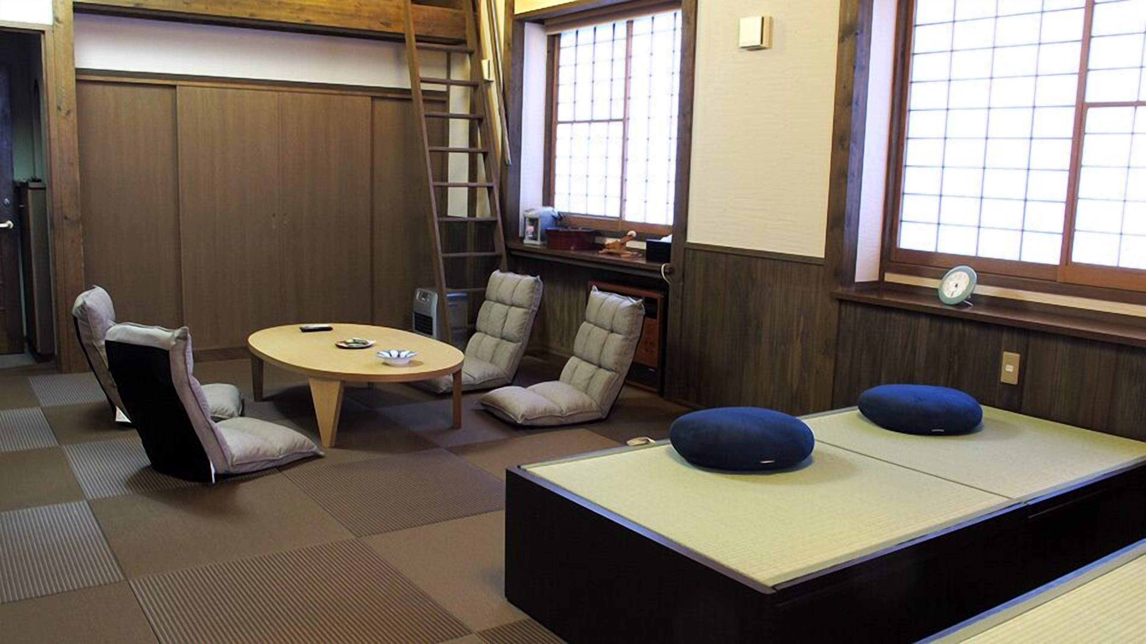 Une pièce avec un sol recouvert de tatamis, une petite table basse, deux chaises et un futon servant de lit.