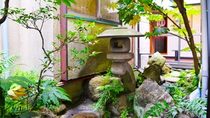 Un farol de piedra tradicional rodeado de exuberante follaje y rocas, frente a una fachada de edificio.
