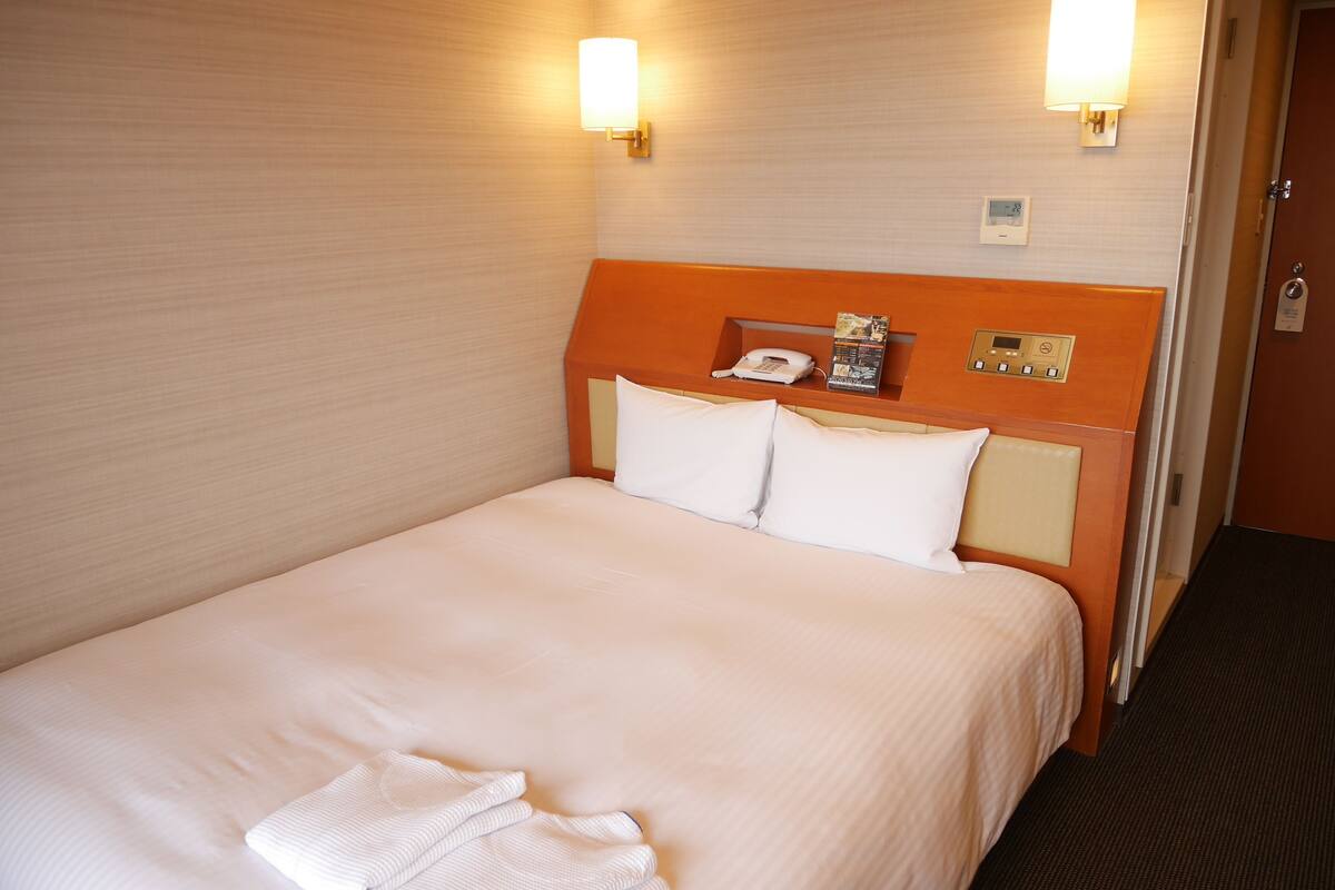 Une chambre d’hôtel avec un lit simple, un téléphone sur la table de chevet, un panneau de commande et deux lampes fixées au mur.