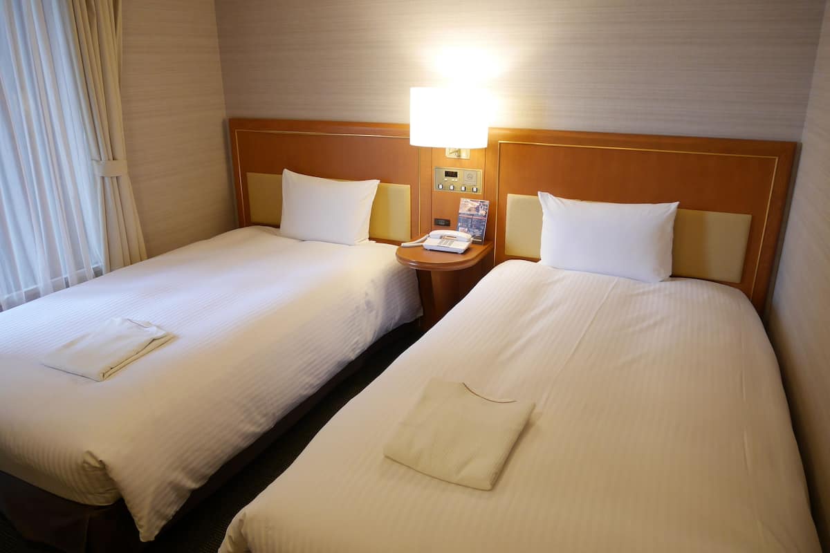 Une chambre d’hôtel avec deux lits, chacun étant équipé d’un oreiller et d’une serviette pliée. On y trouve une table de chevet avec une lampe et un téléphone, ainsi qu’une petite table sur laquelle est posé un menu.