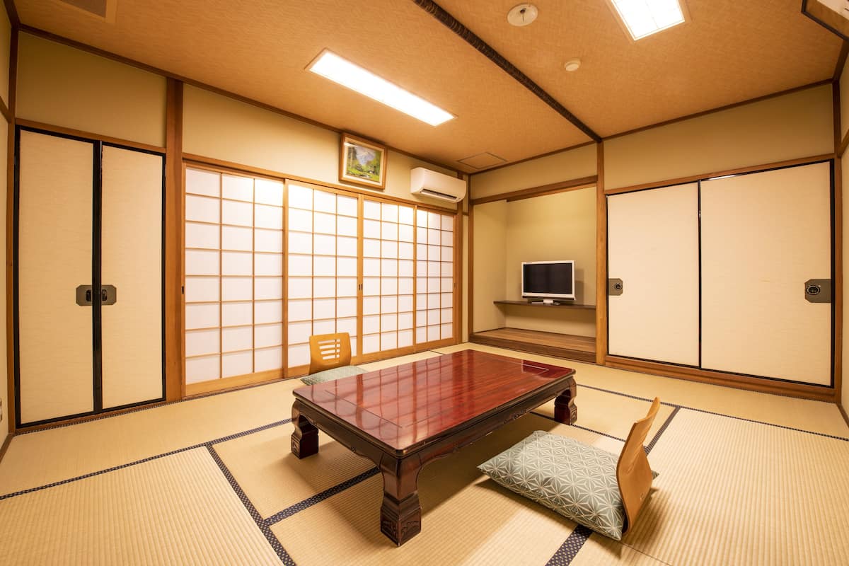 Um quarto japonês tradicional com tatames, uma mesa de madeira baixa, portas deslizantes e uma televisão.