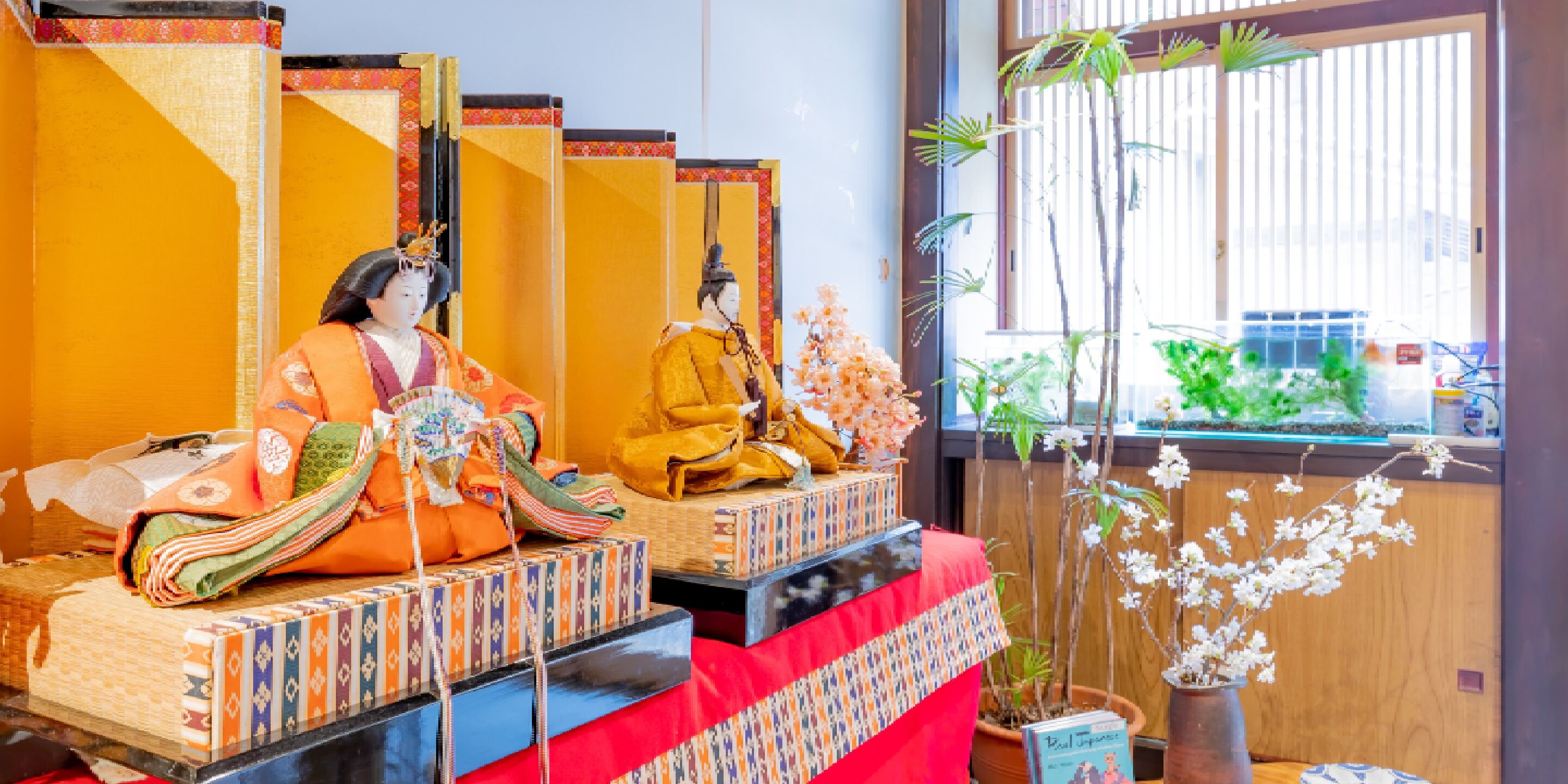 Una habitación japonesa tradicional con tatamis, una mesa baja cubierta con un paño rojo y una ventana con vistas a la vegetación.