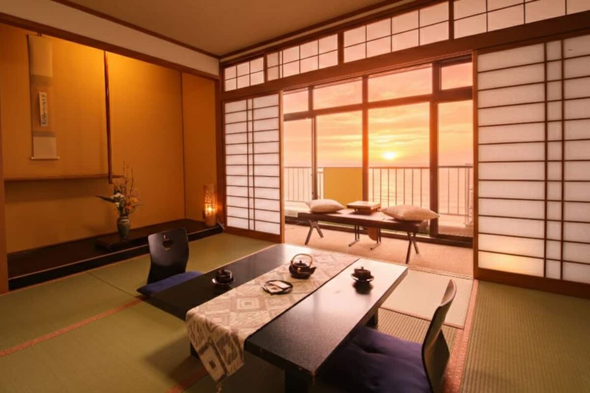 A traditional Japanese room with tatami flooring, a low table with cushions, a sliding door with a view, and a small table with a vase and teapot.