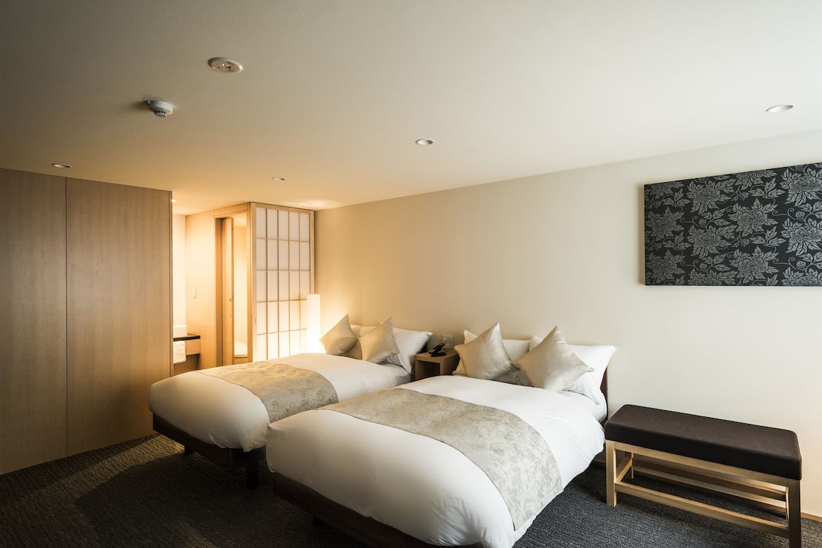 A hotel room with two beds, a bench, a bedside table, a wall-mounted artwork, and a sliding door.