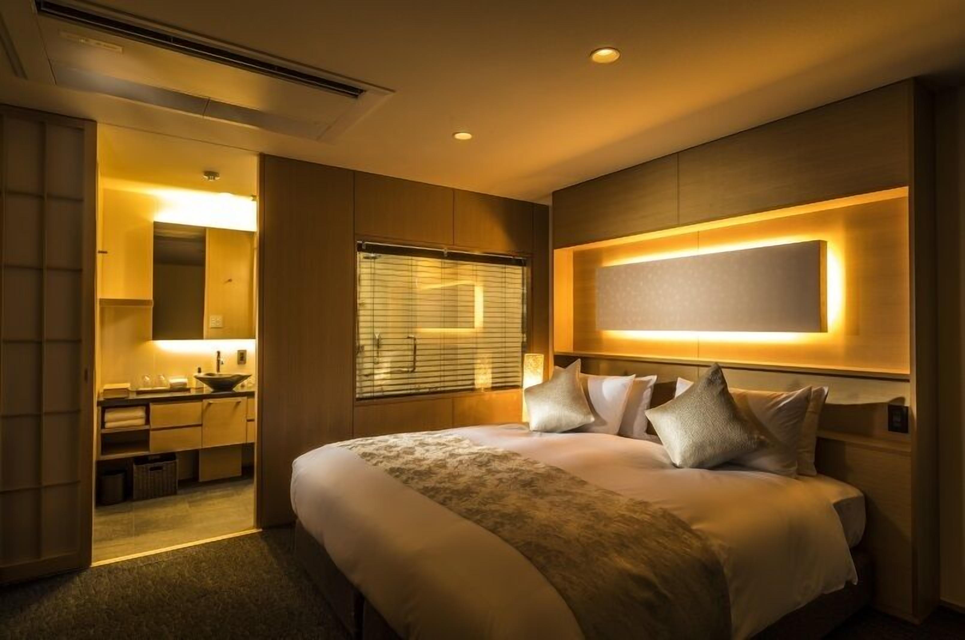 A modern hotel room with a large bed, bedside tables, a built-in TV, and a bathroom with a vanity and sink.