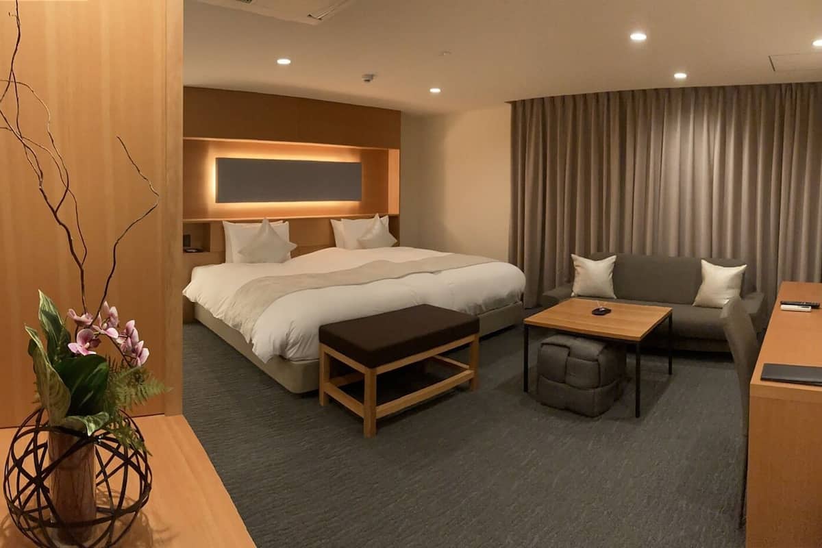 Hotellihuone, jossa on sänky, sohva, pieni pöytä, työpöytä ja koristeellinen kasvi.
