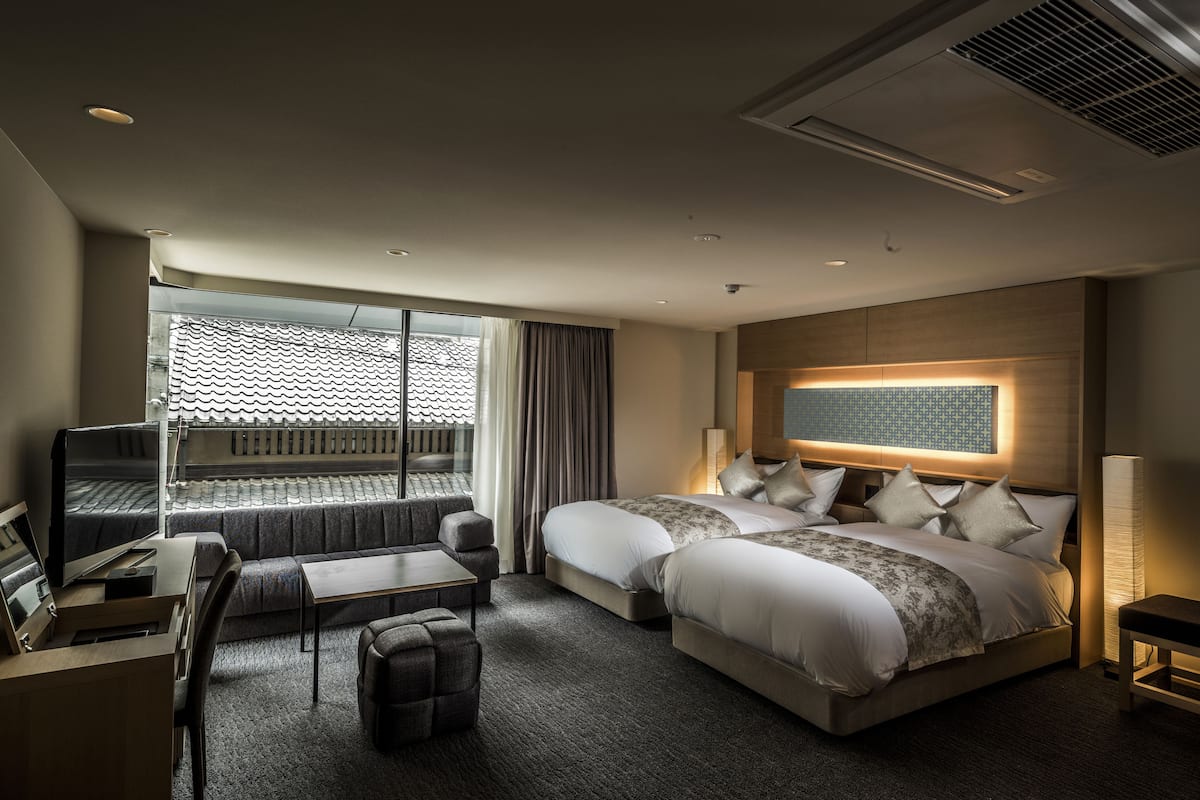 A hotel room with a large bed, a desk with a computer, a seating area, and a window with a view of a tiled roof.