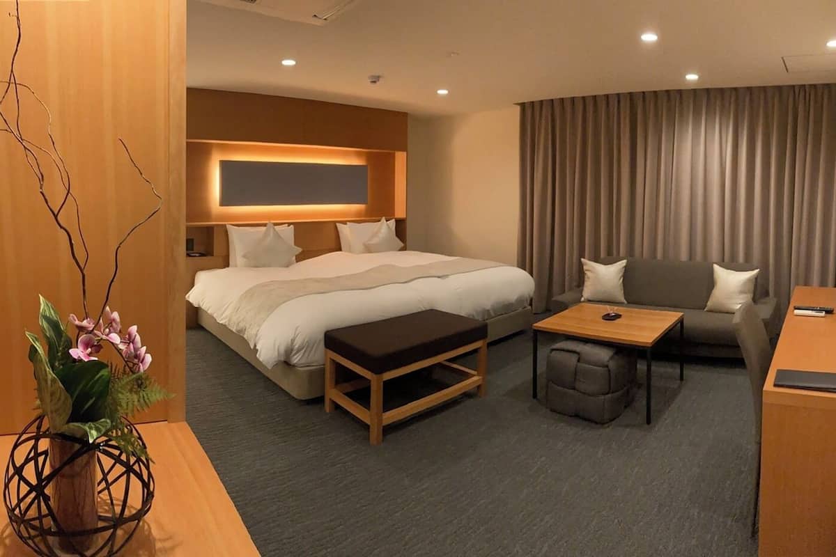 A hotel room with a bed, a sofa, a small table, a desk, and a decorative plant.
