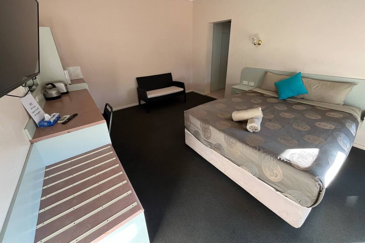 Hotellihuone, jossa on sänky, työpöytä, tuoli, penkki ja seinään kiinnitetty valaisin.
