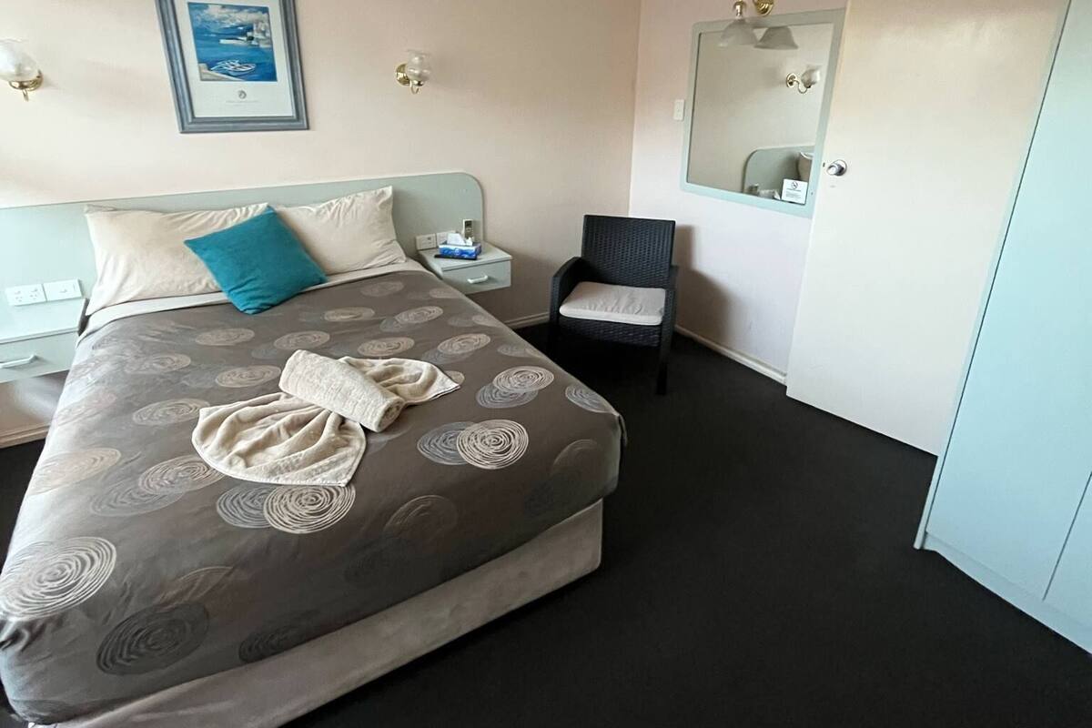 Hotellihuone, jossa on suuri sänky, tuoli, yöpöytä, seinällä oleva kehystetty kuva ja ovi.