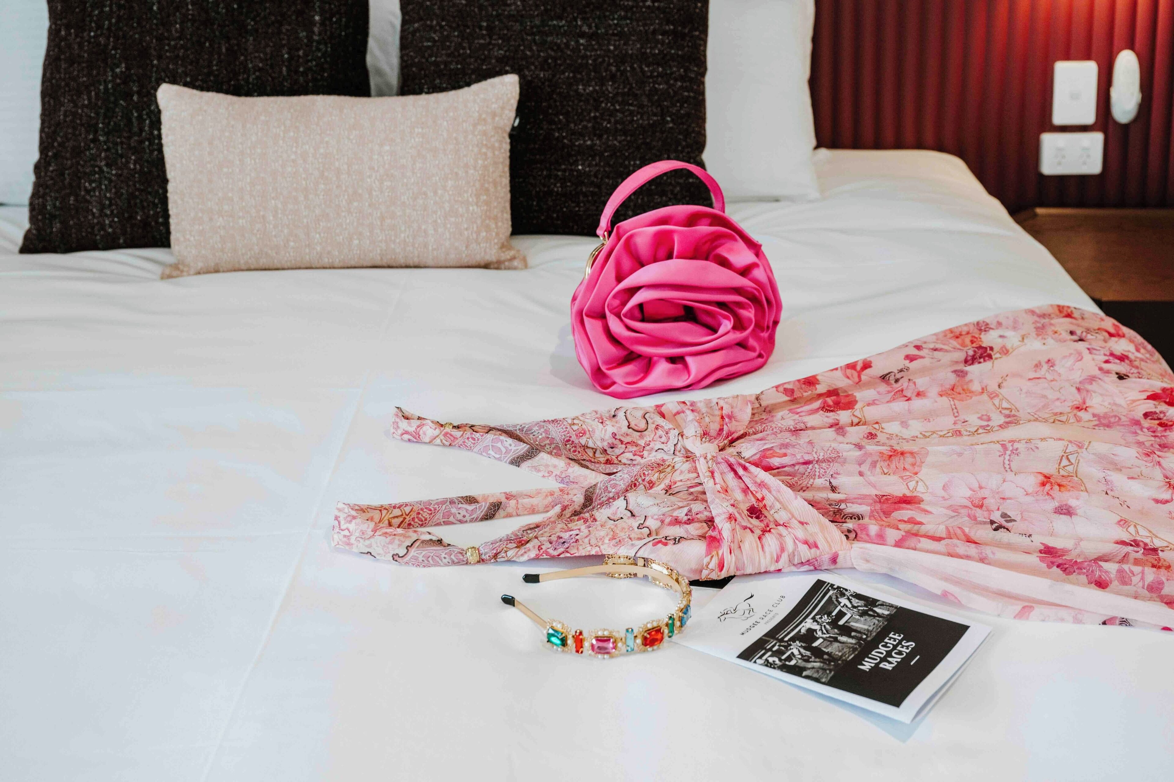 A neatly made bed with a pink floral blanket, a pink handbag, a decorative headband, and a magazine on the bed.