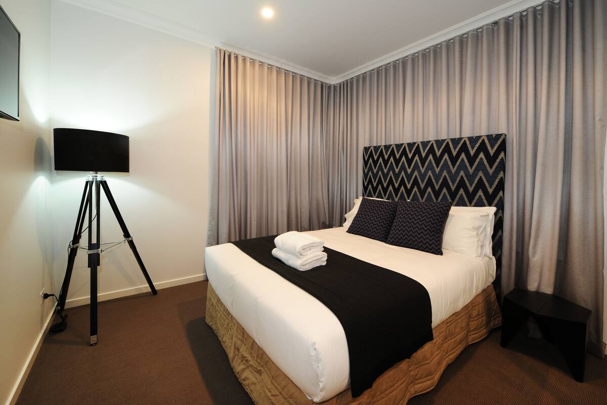A neatly made bed with a black and white headboard, a lamp on a tripod stand, and a small table beside the bed.