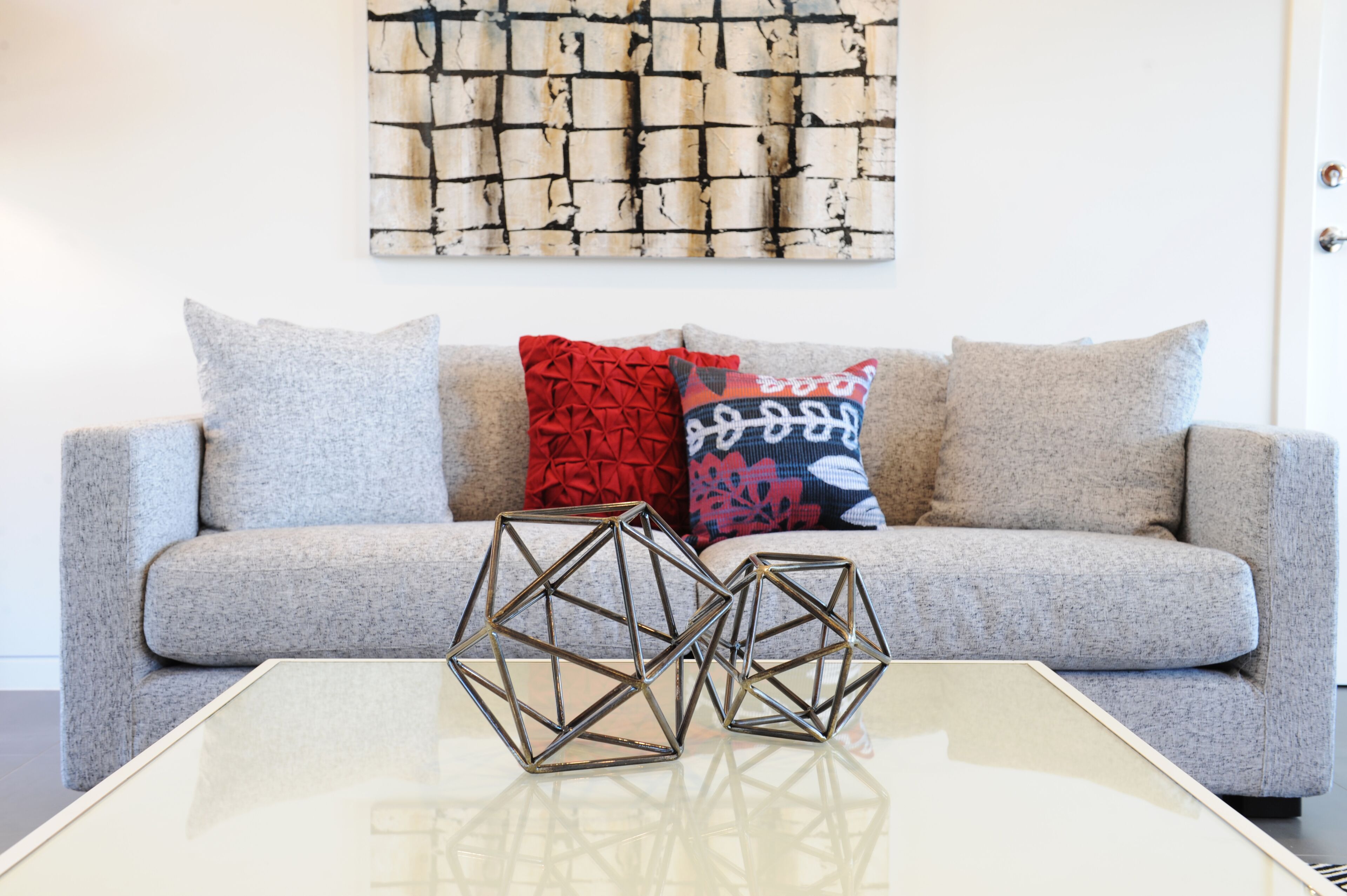 A modern living room with a grey sofa, two geometric metal sculptures on a glass coffee table, and patterned throw pillows.