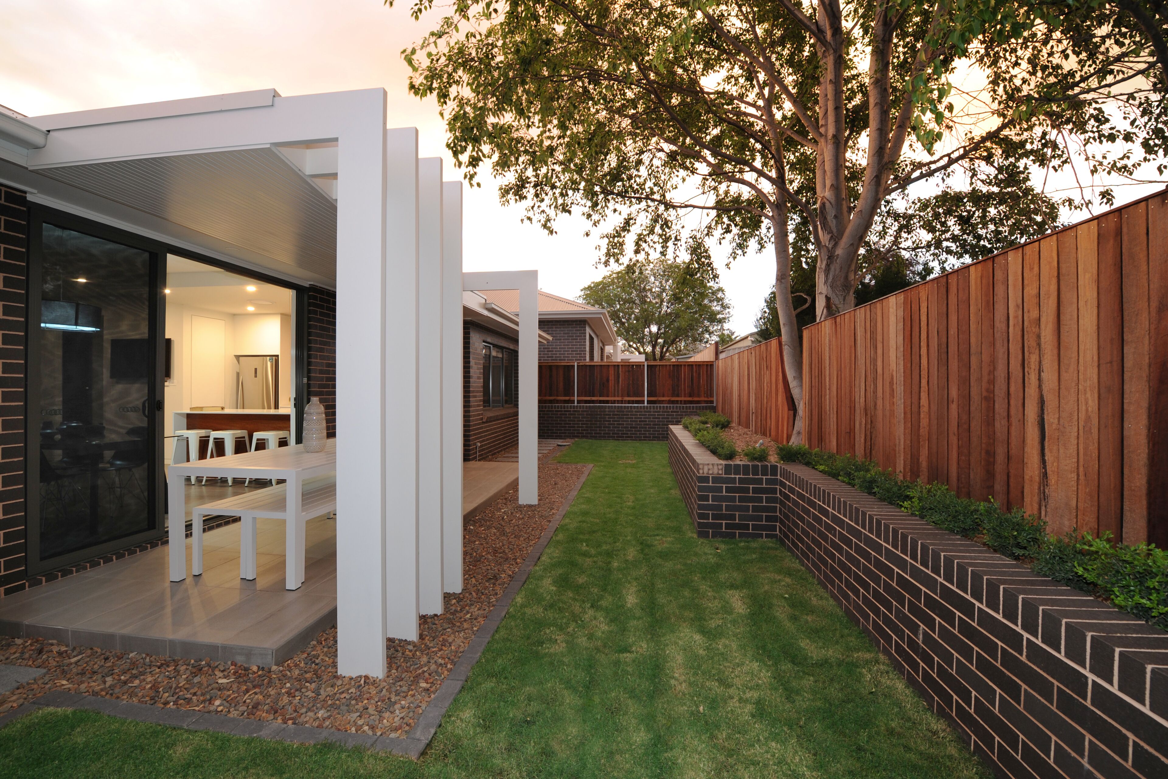 Un patio exterior moderno con una mesa y sillas blancas, una cerca de madera y un césped bien cuidado.