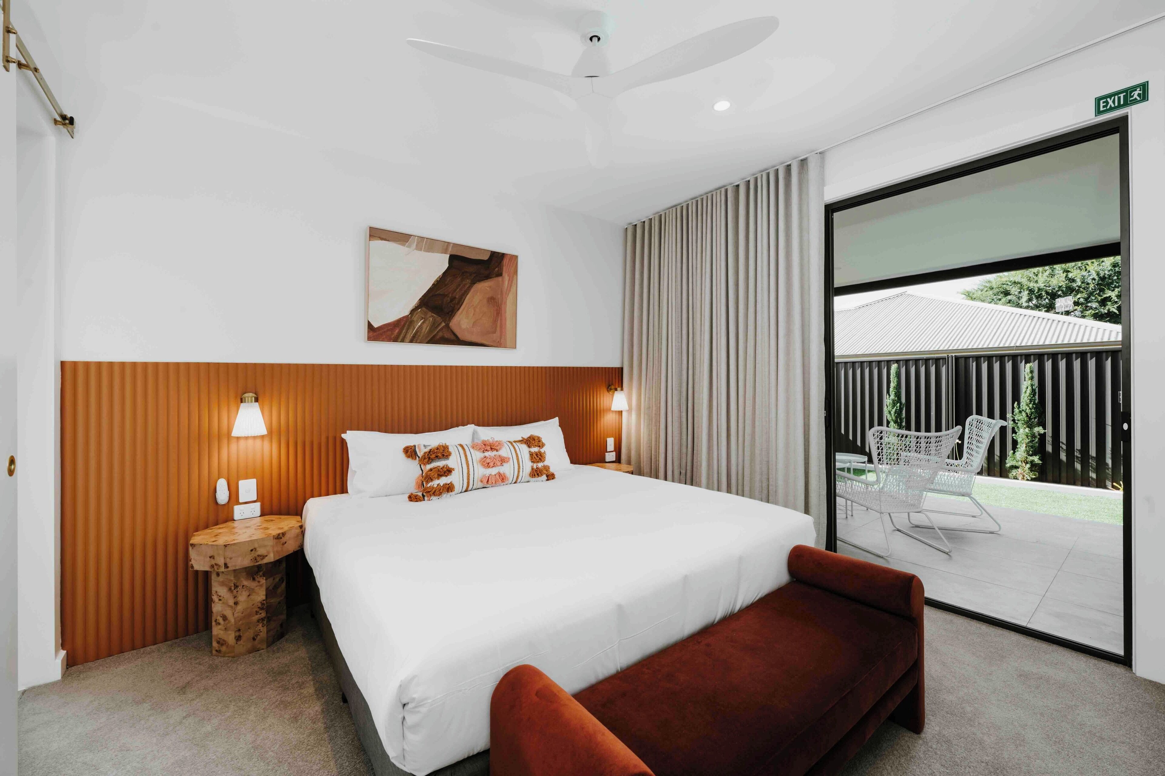 Quarto de hotel moderno com uma cama grande, cabeceira de madeira, mesa de cabeceira com abajur, um banco e uma porta de vidro deslizante para uma área externa com uma cadeira.