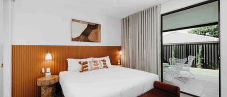 Quarto de hotel moderno com uma cama grande, cabeceira de madeira, mesa de cabeceira com abajur, um banco e uma porta de vidro deslizante para uma área externa com uma cadeira.