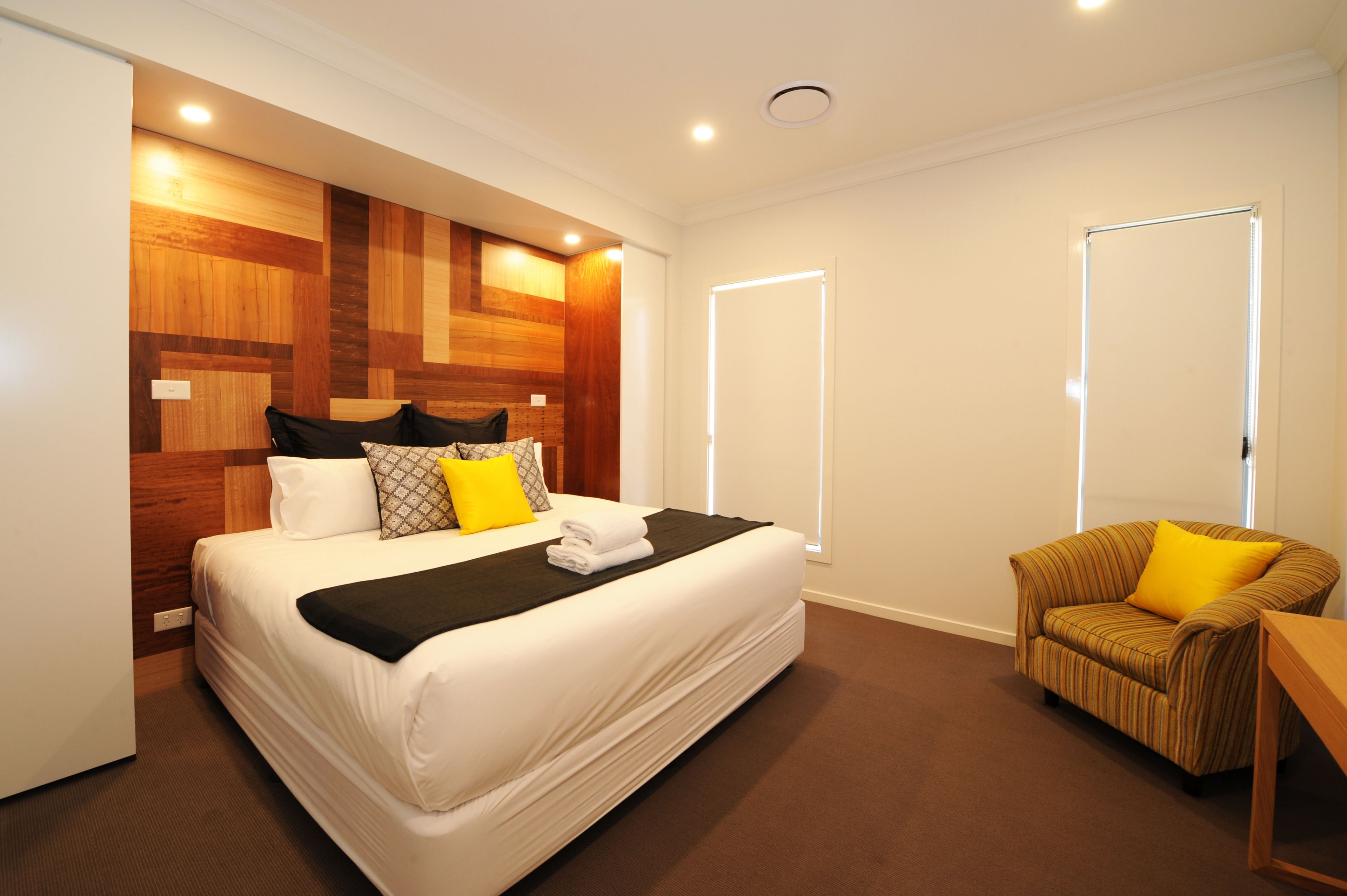 A hotel room with a large bed, a wooden headboard, a chair with a yellow pillow, and a bedside table.