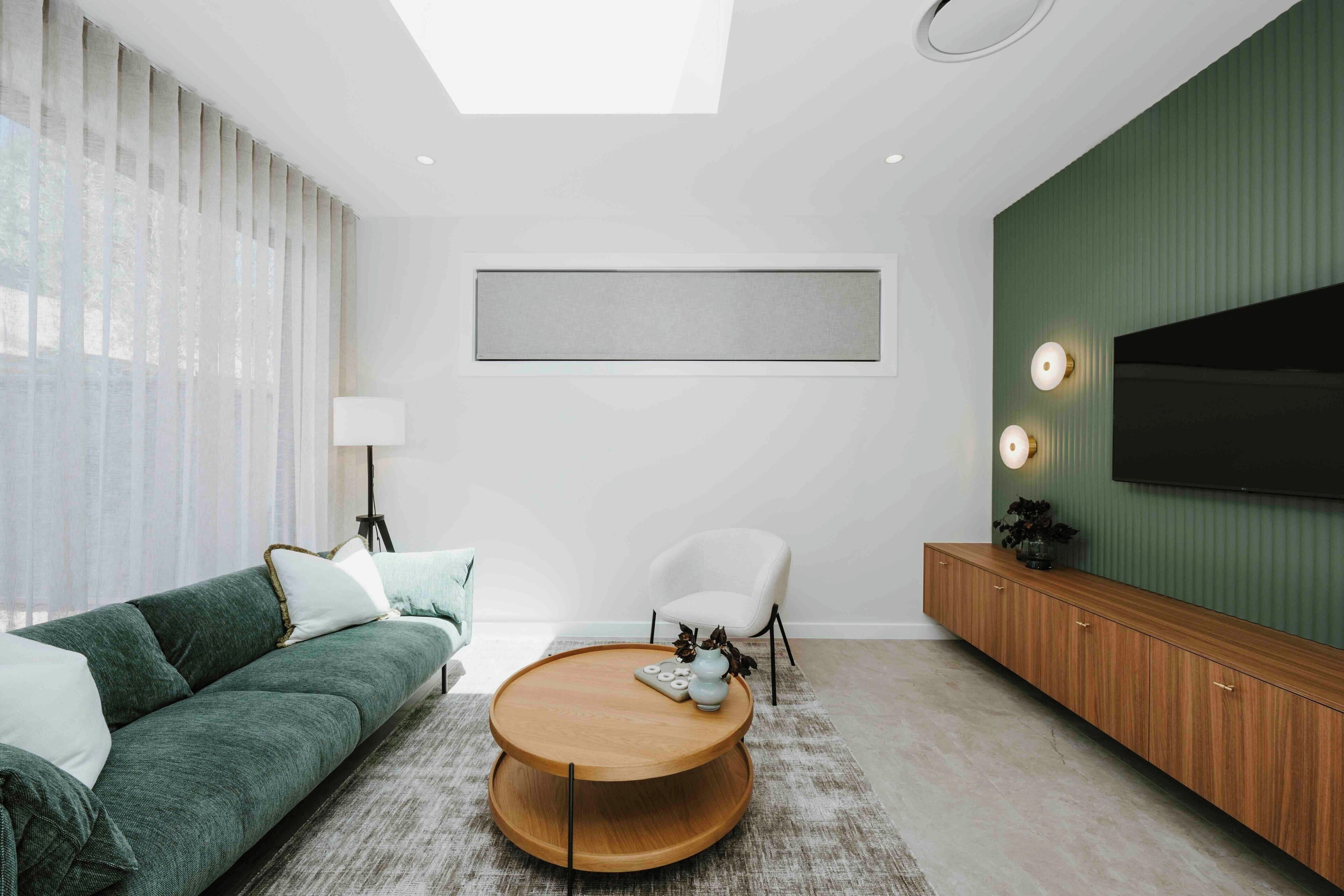 A modern living room with a green sofa, a round wooden coffee table, a white armchair, a TV mounted on a green wall, and a floor lamp.
