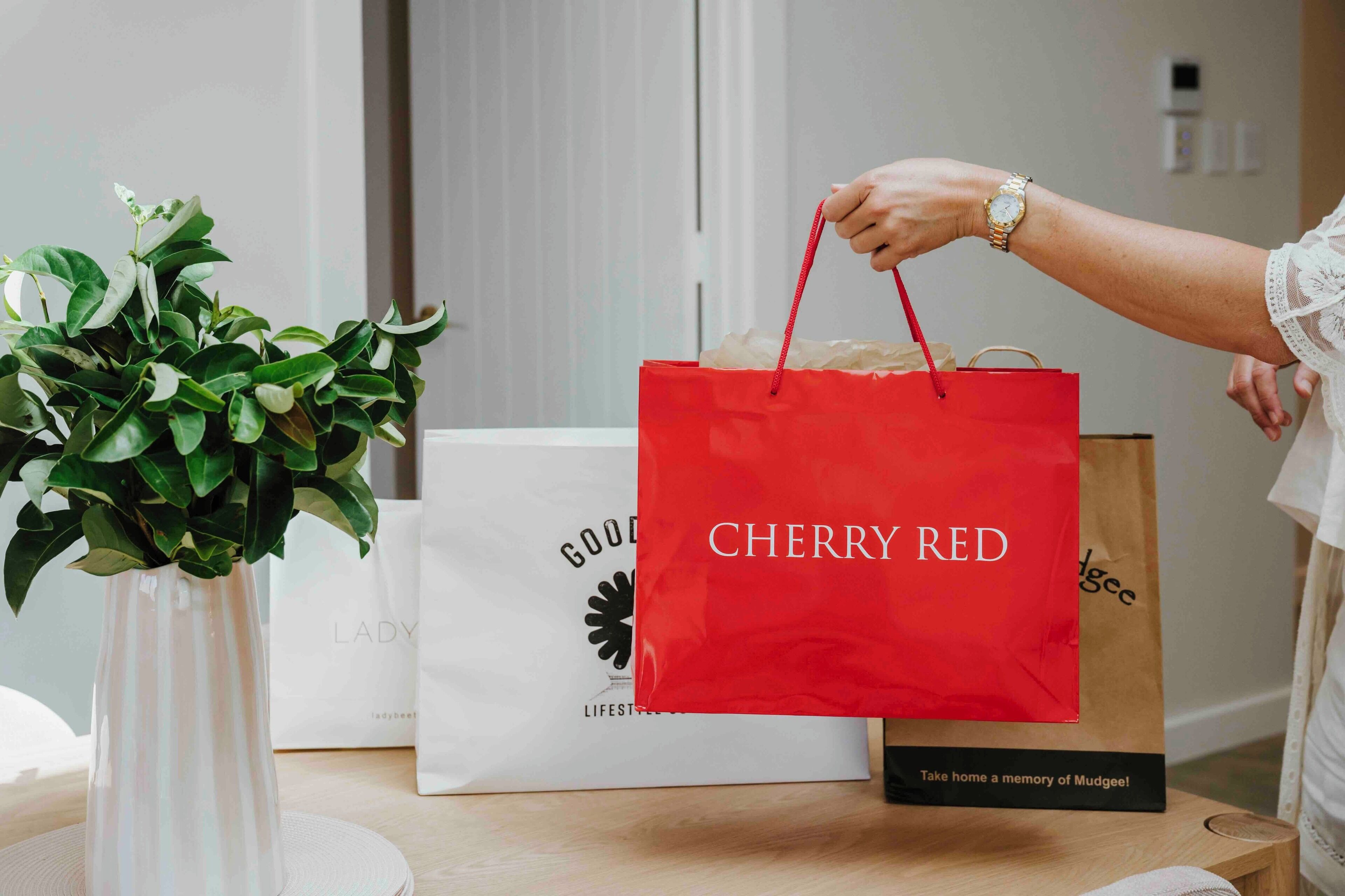 A hand holding a red shopping bag with "CHERRY RED" written on it, placed on a table with a white vase and a white bag with "LADY" written on it.