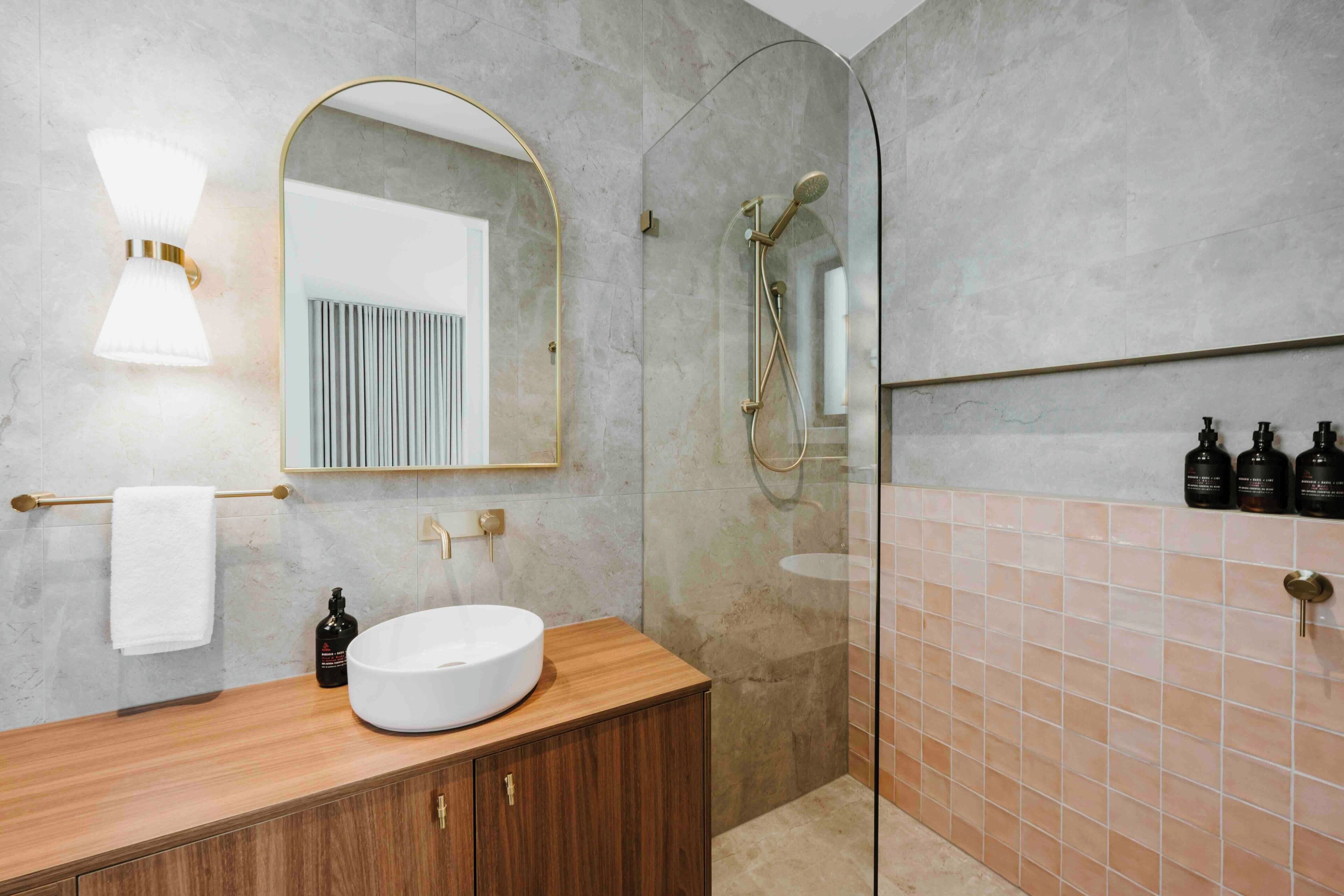 A modern bathroom with a wooden vanity, a round sink, a mirror with a gold frame, a glass-enclosed shower, and a tiled wall.