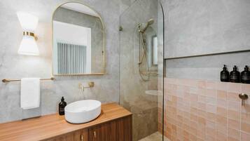 A modern bathroom with a wooden vanity, a round sink, a mirror with a gold frame, a glass-enclosed shower, and a tiled wall.