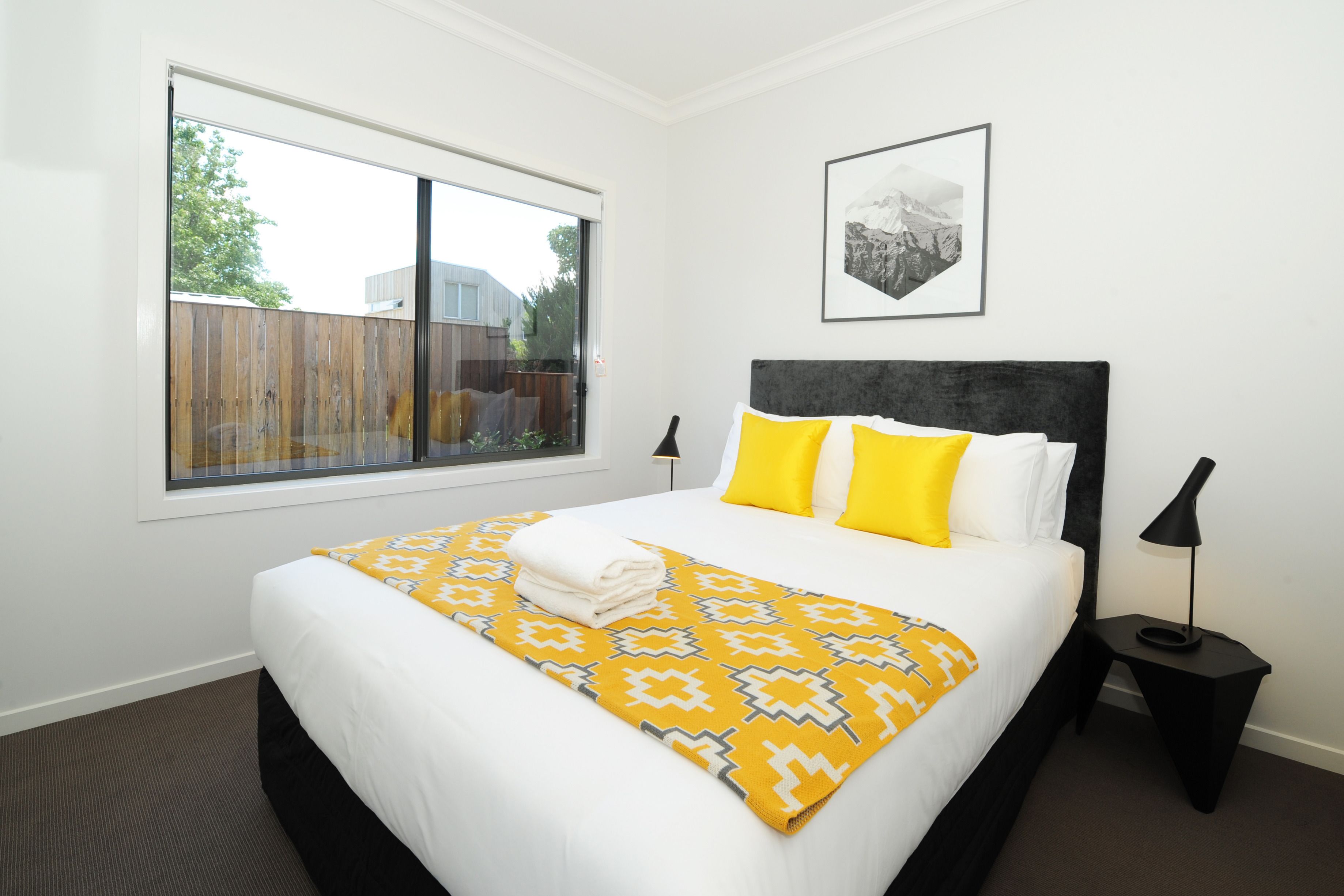 Habitación de hotel con una cama grande, ventana con vistas a una valla, un cuadro enmarcado en la pared y una mesita con una lámpara.