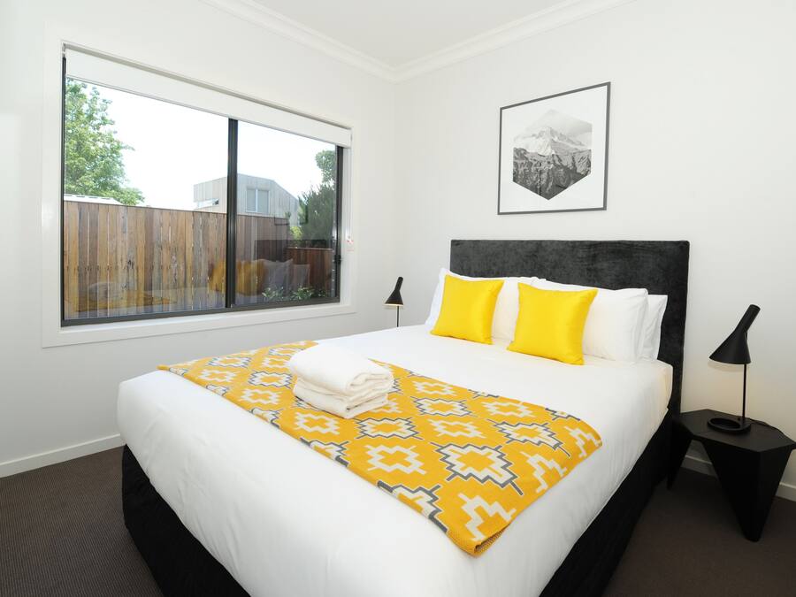 Habitación de hotel con una cama grande, ventana con vistas a una valla, un cuadro enmarcado en la pared y una mesita con una lámpara.