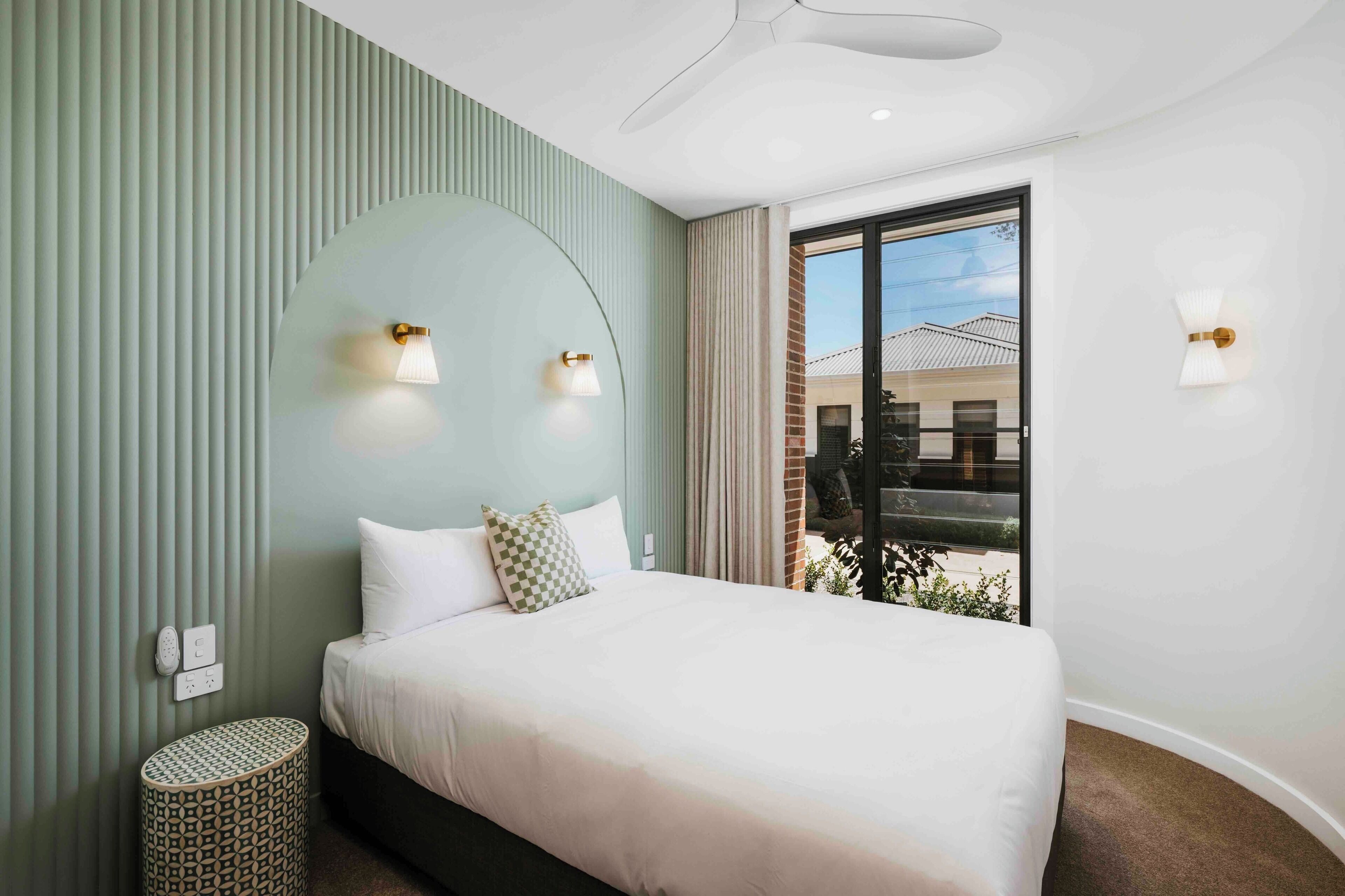 Une chambre d’hôtel moderne, équipée d’un lit, de deux lampes fixées au mur, d’un ventilateur de plafond et offrant une vue sur l’extérieur.
