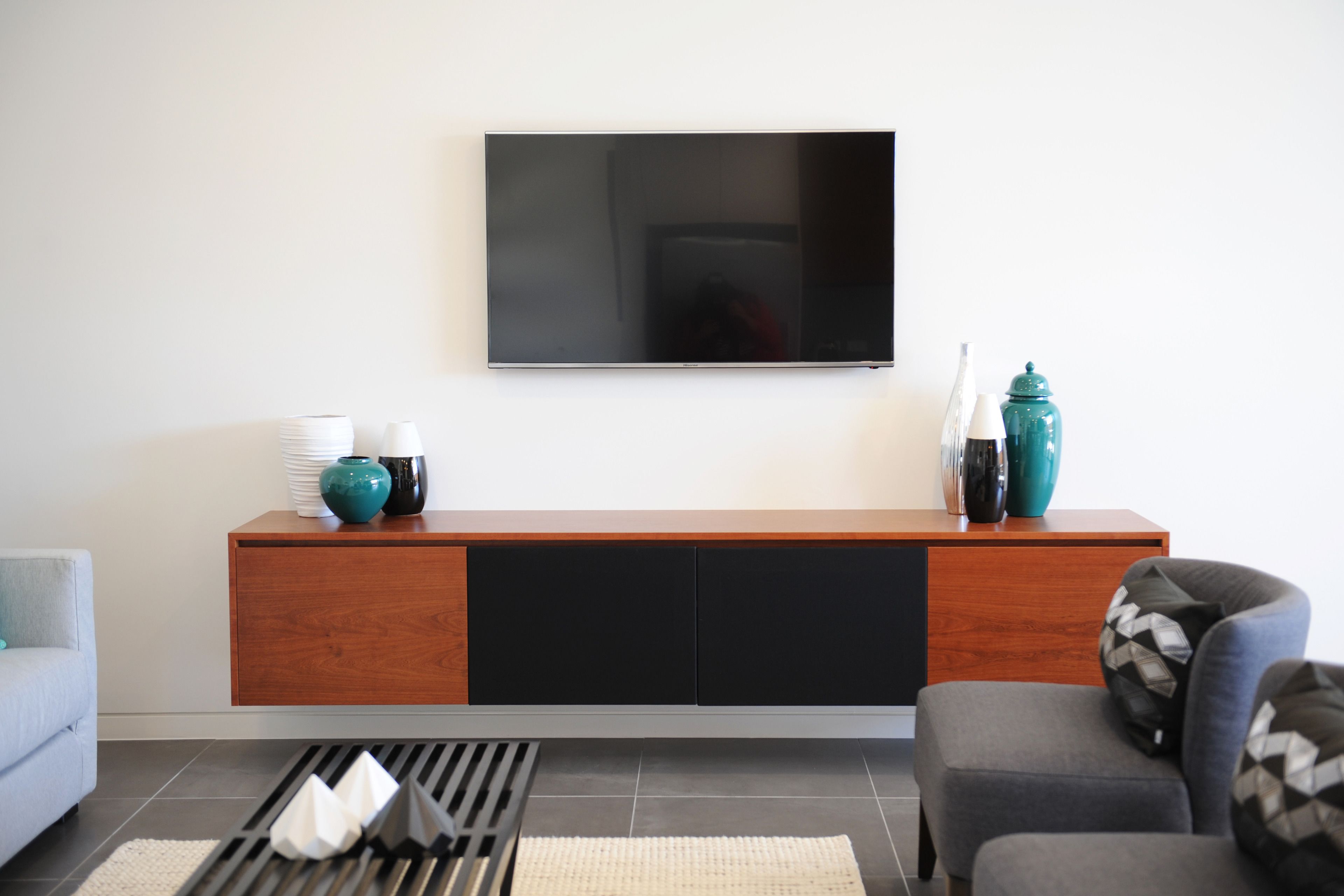 Una sala de estar moderna con un televisor de pantalla plana montado en la pared, una consola de madera con objetos decorativos, un sofá gris y un sillón estampado.