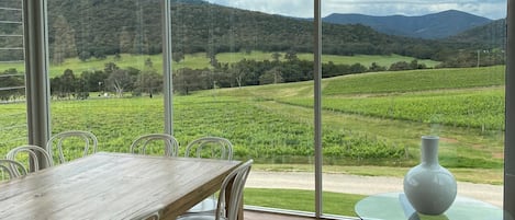 Un espace repas comprenant une grande table et des chaises, une table en verre, et offrant une vue sur un vignoble et des montagnes grâce à de grandes fenêtres.