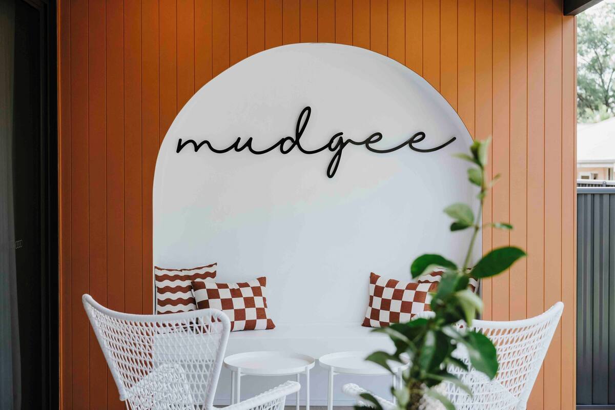 A cozy outdoor seating area with white wicker chairs and a table, set against a wooden wall with a 'mudgee' sign above.