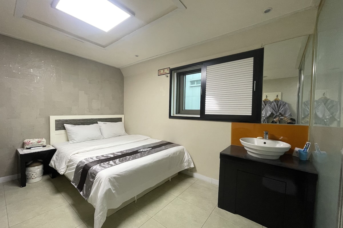 A hotel room with a bed, a nightstand, a bathroom with a sink and mirror, and a window with blinds.
