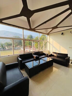 A modern lounge with black leather sofas and glass coffee tables, large windows offering a view of a playground and hills. - Ritumbraha Hotel & Resort (Alwar)