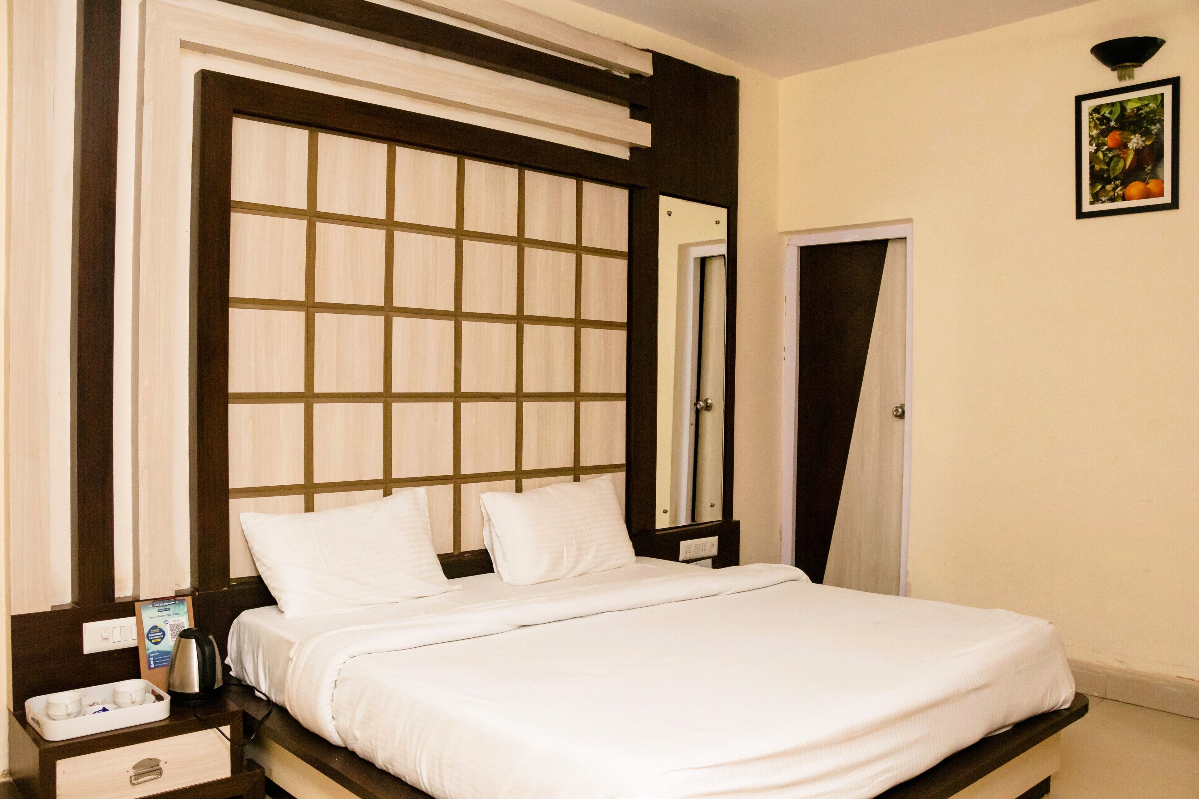 Een hotelkamer met een groot bed, een nachtkastje met een waterkoker, een klein lade en een lijst met een foto aan de muur.