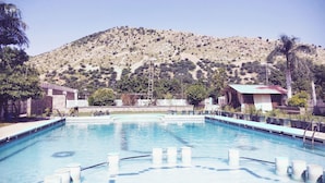 A swimming pool with clear blue water, surrounded by a white deck and palm trees. In the background, there is a hill with sparse vegetation and a few buildings.