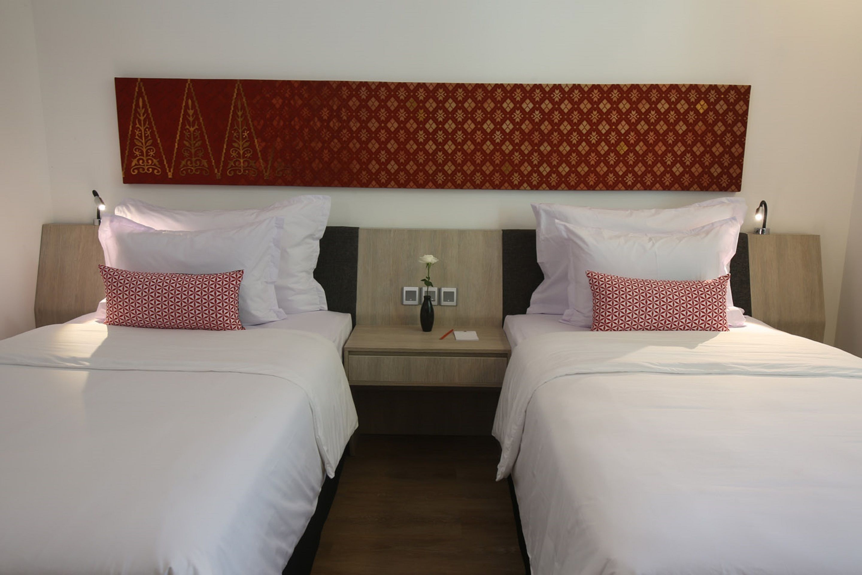 A hotel room with two beds, each with white bedding and red patterned pillows. There is a wooden headboard with a decorative red panel above the beds. A small table with a vase of flowers and a few items is between the beds.