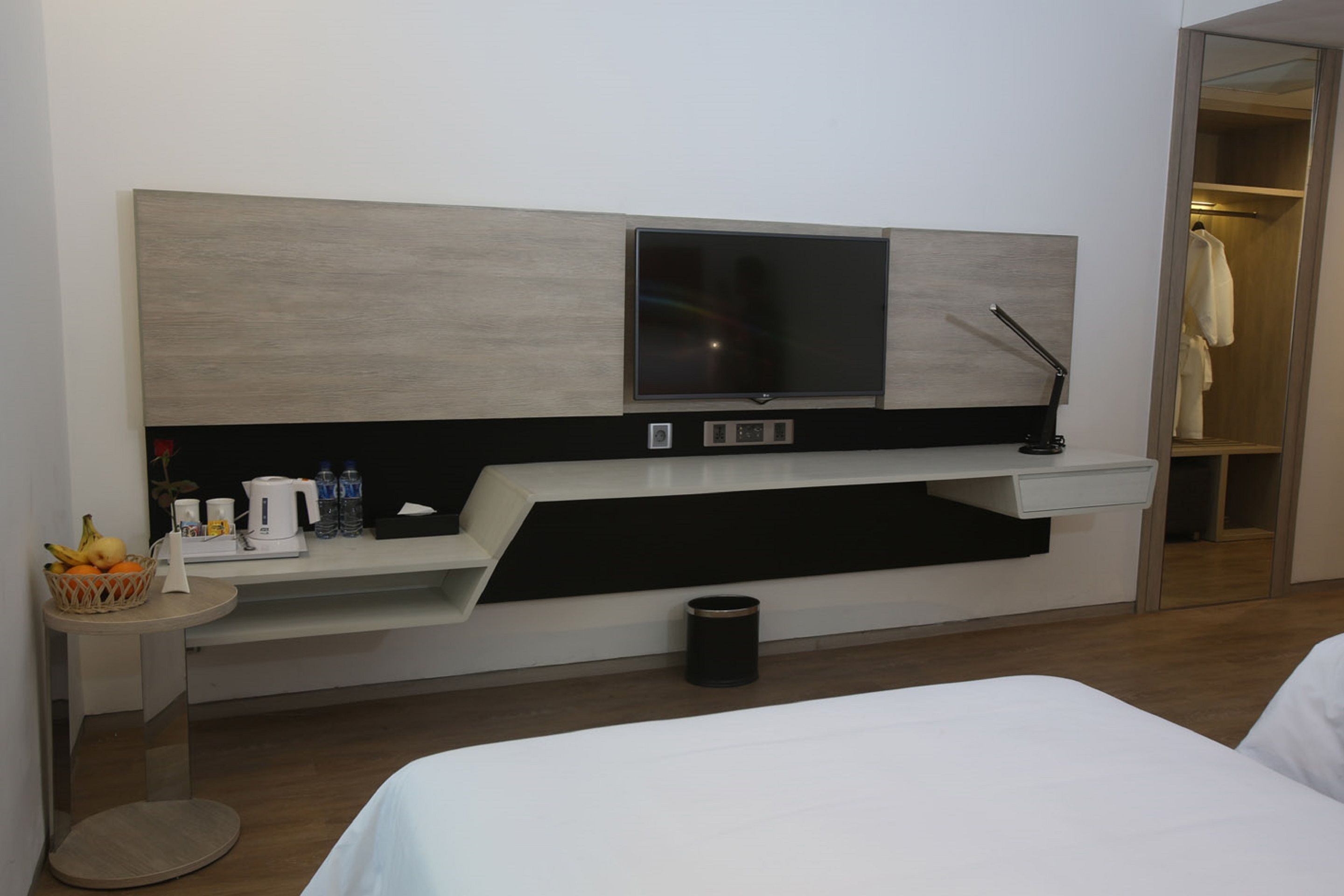 Ein modernes Hotelzimmer mit Flachbildfernseher an einer Holzvertäfelung, einem kleinen Nachttisch mit Obstkorb, einer Kaffeemaschine und einem Papierkorb.